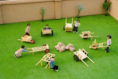 Bulk preschool gross motor equipment featuring a group of kids playing with an 8-piece kindergarten outdoor play wooden transport and logistics cart set on artificial grass.