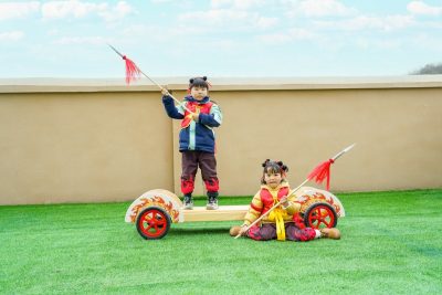 Bulk preschool gross motor equipment featuring children on a customized kindergarten outdoor play wooden rowing chariot with safety spears on artificial turf.